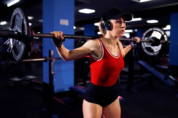 Bela mulher sexy desportiva em boné e fones de ouvido fazendo agachamento treino no ginásio — Fotografia de Stock