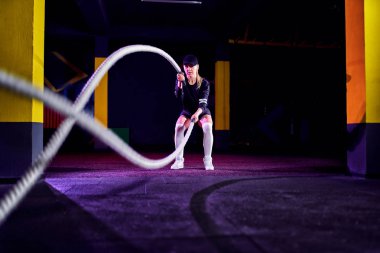 Spor salonundaki antrenman iplerini beden eğitimi için kullanan bir kadın. Sporcu çapraz spor salonunda savaş halatlarıyla çalışıyor.