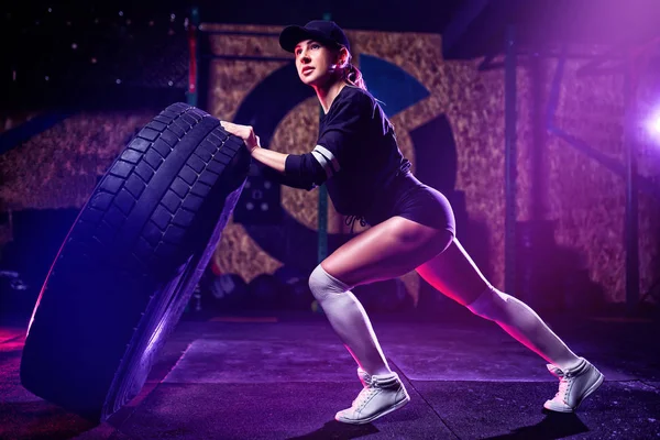 Atractiva atleta en forma de mujer haciendo ejercicio con un neumático enorme, girando y volteando en el gimnasio. Mujer en forma de cruz haciendo ejercicio con neumático grande — Foto de Stock