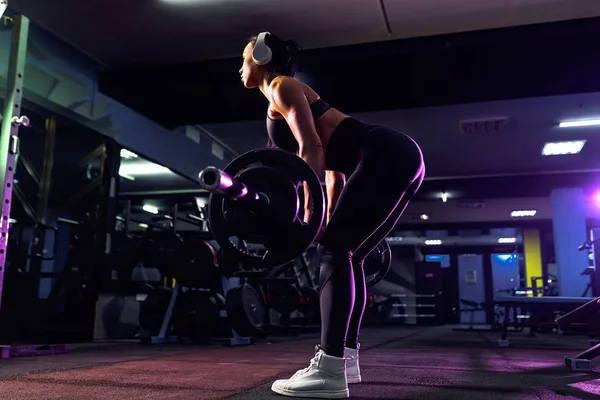 Aantrekkelijke pasvorm sexy vrouw in draadloze hoofdtelefoon luisteren muziek en doen hurken met een lange halter in de sportschool. Vrouwentraining terug — Stockfoto