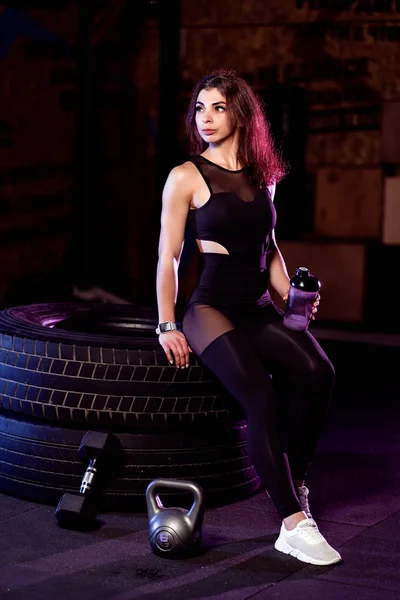 Приваблива жінка з шейкером позує на велику шину в спортзалі — стокове фото