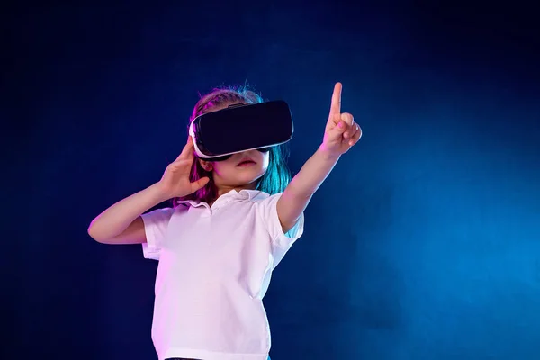 Menina 7 anos. experimentando jogo de fone de ouvido VR em fundo colorido. Criança apontando o dedo ao usar um gadget de jogo para realidade virtual . — Fotografia de Stock