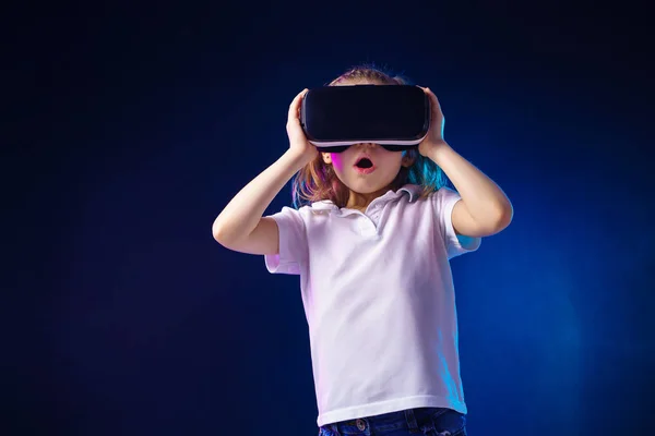 Menina 7 anos. experimentando jogo de fone de ouvido VR em fundo colorido. Emoções surpresas em seu rosto.Criança usando um gadget de jogo para realidade virtual . — Fotografia de Stock