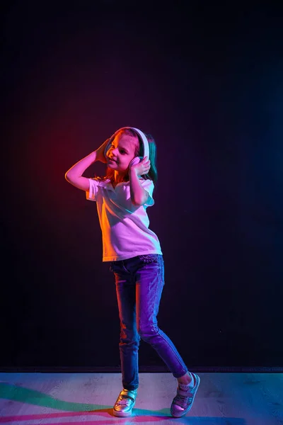Girl listening to music in headphones on dark colorful background. Neon light. Dancing girl. Happy small girl dancing to music. Cute child enjoying happy dance music.