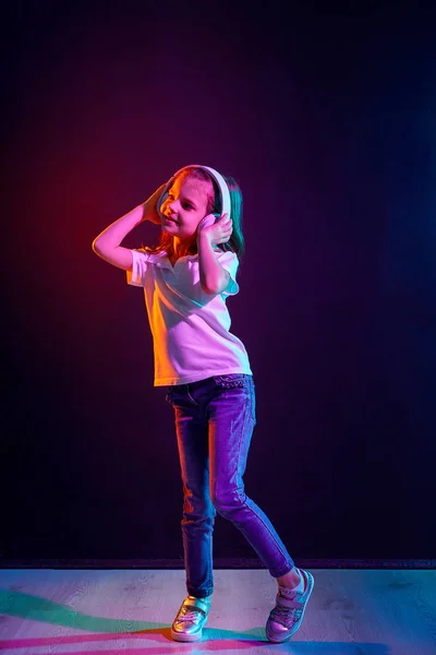 Chica escuchando música en auriculares sobre un fondo oscuro y colorido. Luz de neón. Bailarina. Feliz niña bailando música. Lindo niño disfrutando de música de baile feliz . — Foto de Stock