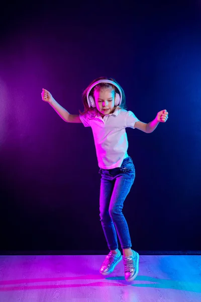 Chica escuchando música en auriculares sobre un fondo oscuro y colorido. Bailarina. Feliz niña bailando música. Lindo niño disfrutando de música de baile feliz . — Foto de Stock