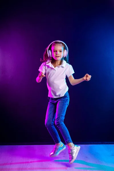暗いカラフルな背景にヘッドフォンで音楽を聴く女の子.踊ってる女の子音楽に踊る幸せな小さな女の子。幸せなダンスミュージックを楽しむかわいい子供. — ストック写真