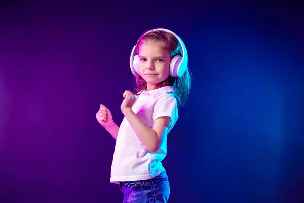 Chica escuchando música en auriculares sobre un fondo oscuro y colorido. Lindo niño disfrutando de música de baile feliz, mirando a la cámara y la sonrisa posando en la pared de fondo del estudio — Foto de Stock