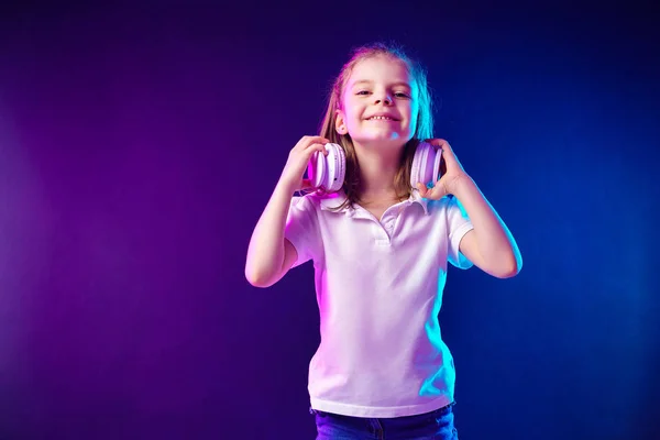 Lány hallgató-hoz zene-ban fejhallgató-ra sötét színdús háttér. Aranyos gyerek élvezi boldog tánczene, néztem kamerát és mosolyt jelentő stúdió háttérfal — Stock Fotó