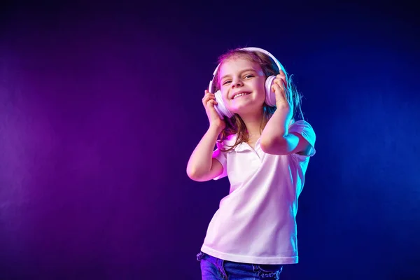 Chica escuchando música en auriculares sobre un fondo oscuro y colorido. Bailarina. Feliz niña bailando música. Lindo niño disfrutando de música de baile feliz . — Foto de Stock