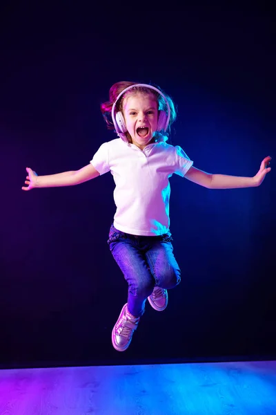 Chica escuchando música en auriculares sobre un fondo oscuro y colorido. Bailarina. Feliz niña bailando música. Lindo niño disfrutando de música de baile feliz . — Foto de Stock