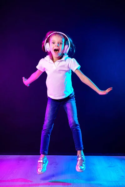 Κορίτσι ακούγοντας μουσική στα ακουστικά σε σκούρο πολύχρωμο φόντο. Το κορίτσι που χορεύει. Ευτυχισμένο μικρό κορίτσι που χορεύει στη μουσική. Χαριτωμένο παιδί απολαμβάνοντας χαρούμενη μουσική χορού. — Φωτογραφία Αρχείου