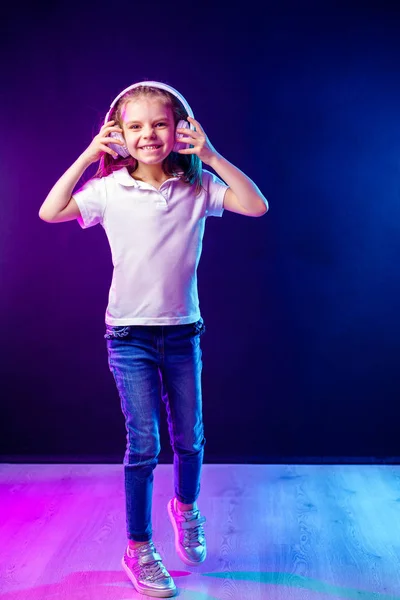 Chica escuchando música en auriculares sobre un fondo oscuro y colorido. Bailarina. Feliz niña bailando música. Lindo niño disfrutando de música de baile feliz . — Foto de Stock