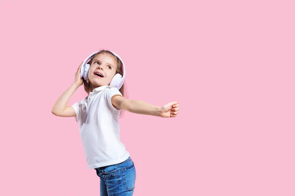 Chica escuchando música en auriculares sobre fondo rosa. Lindo niño disfrutando de música de baile feliz, ojos cerrados y sonrisa posando en la pared de fondo del estudio — Foto de Stock