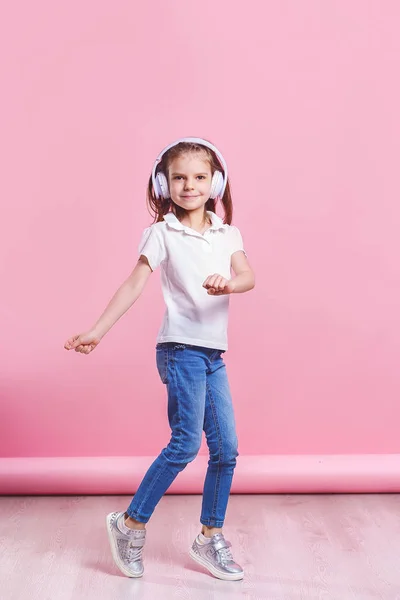 ヘッドフォンで音楽を聴く女の子は、ピンクの背景にダンス。幸せなダンスミュージックを楽しむかわいい子供、目を閉じて、スタジオの背景の壁にポーズ笑顔 — ストック写真