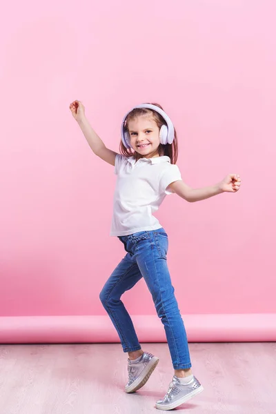 Chica escuchando música en auriculares un baile sobre fondo rosa. Lindo niño disfrutando de música de baile feliz, ojos cerrados y sonrisa posando en la pared de fondo del estudio — Foto de Stock