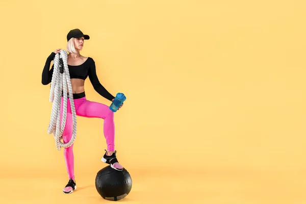 Attractive woman in pink and black sportswear with heavy ropes and shaker on yellow background. — Stock Photo, Image