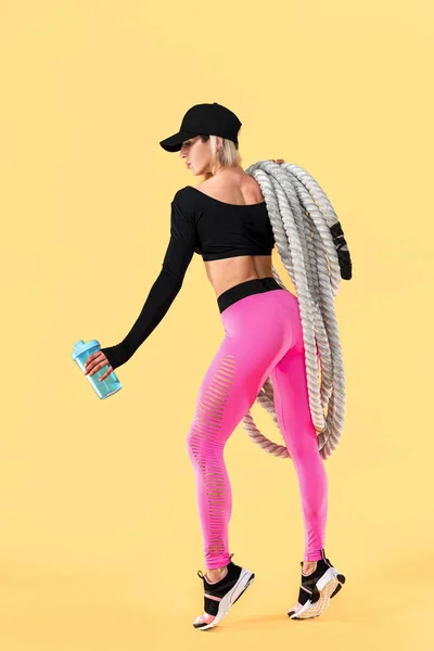 Visão traseira da mulher atraente em roupas esportivas rosa e preto com cordas pesadas em seus ombros e segurando agitador no fundo amarelo. Força e motivação. Mulher desportiva trabalhando com cordas pesadas — Fotografia de Stock
