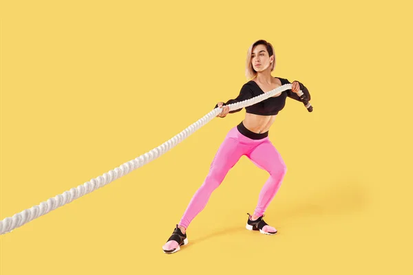 Una mujer fuerte tirando de la cuerda. Foto de mujer atractiva deportiva en ropa deportiva rosa y negra aislada sobre fondo amarillo. Fuerza y motivación —  Fotos de Stock