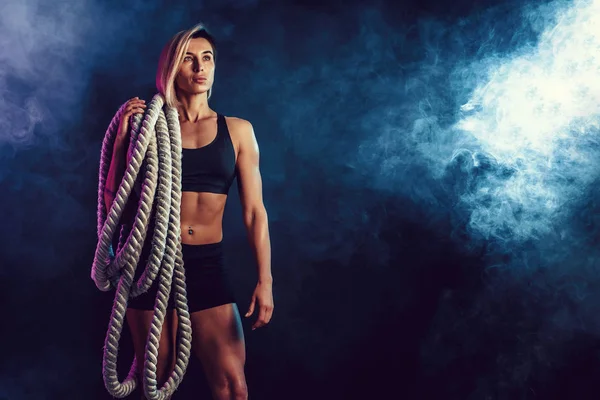 Mujer atractiva en ropa deportiva negra con pesadas cuerdas en los hombros sobre fondo oscuro. Fuerza y motivación. Mujer deportiva trabajando con cuerdas pesadas. Humo. . — Foto de Stock