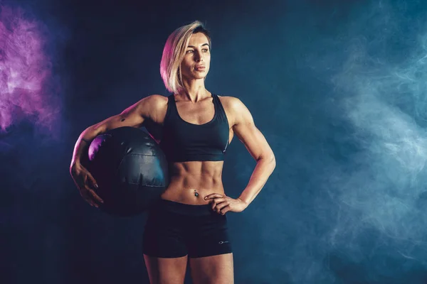 Sporty blonde woman in fashionable sportswear posing with medicine ball. Photo of muscular woman on dark background with smoke. — Stock Photo, Image