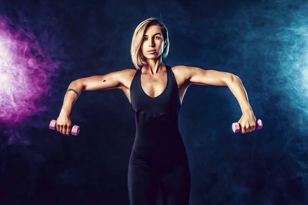 Sporty blonde woman in fashionable sportswear does the exercises with dumbbells. Photo of muscular woman on dark background with smoke. — Stock Photo, Image
