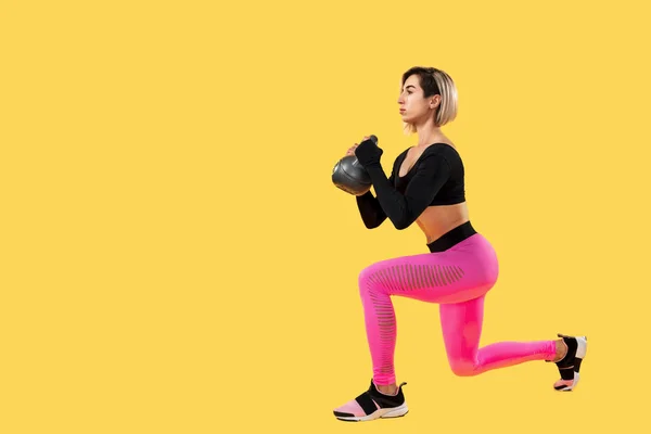 Fitness woman in fashionable pink and black sportswear work out with kettlebell on yellow background. — Stock Photo, Image