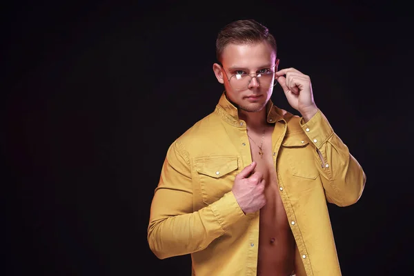 Positive young sexy male looking at camera and adjusting trendy yellow shirt while standing under colorful light during party — Stock Photo, Image