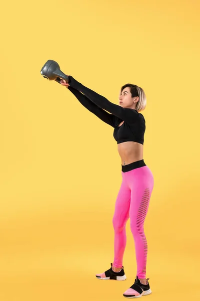 Fitness kobieta w modnej różowej i czarnej odzieży sportowej ćwiczyć z kettlebell na żółtym tle. — Zdjęcie stockowe