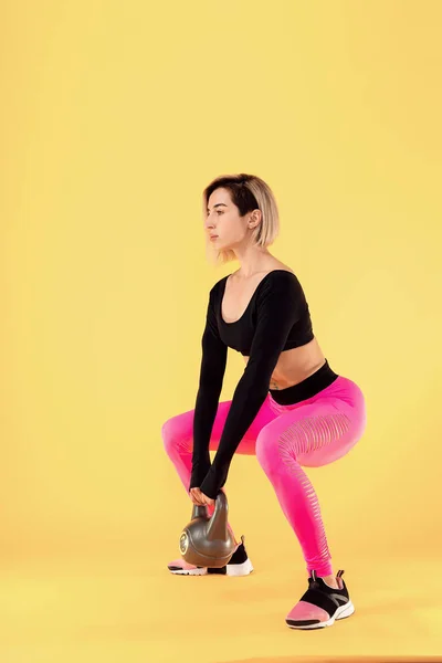 Mujer fitness en ropa deportiva rosa y negro de moda trabajar con kettlebell sobre fondo amarillo . —  Fotos de Stock