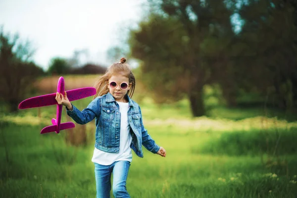 Niña, juega con un avión de juguete en el aire en el parque. Niño lanza un avión de juguete. Hermosa niña, corre sobre la hierba y lanza un avión de juguete rosa — Foto de Stock