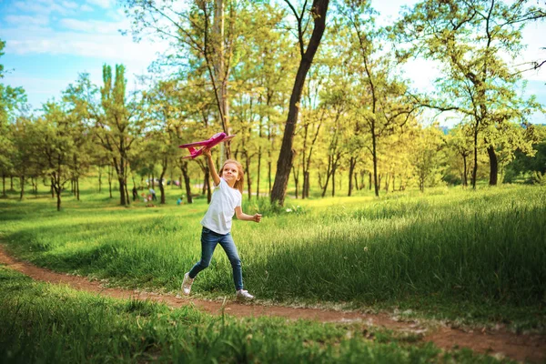 Menina, brinca com um avião de brinquedo no ar no parque. Criança lança um avião de brinquedo. Menina bonita, correr na grama e lança um avião de brinquedo rosa — Fotografia de Stock