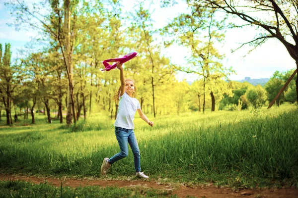 Κοριτσάκι, παίξε με ένα αεροπλανάκι στον αέρα στο πάρκο. Το παιδί εγκαινιάζει ένα παιχνίδι. Όμορφο κοριτσάκι, τρέχει στο γρασίδι και εγκαινιάζει ένα ροζ αεροπλάνο παιχνίδι — Φωτογραφία Αρχείου