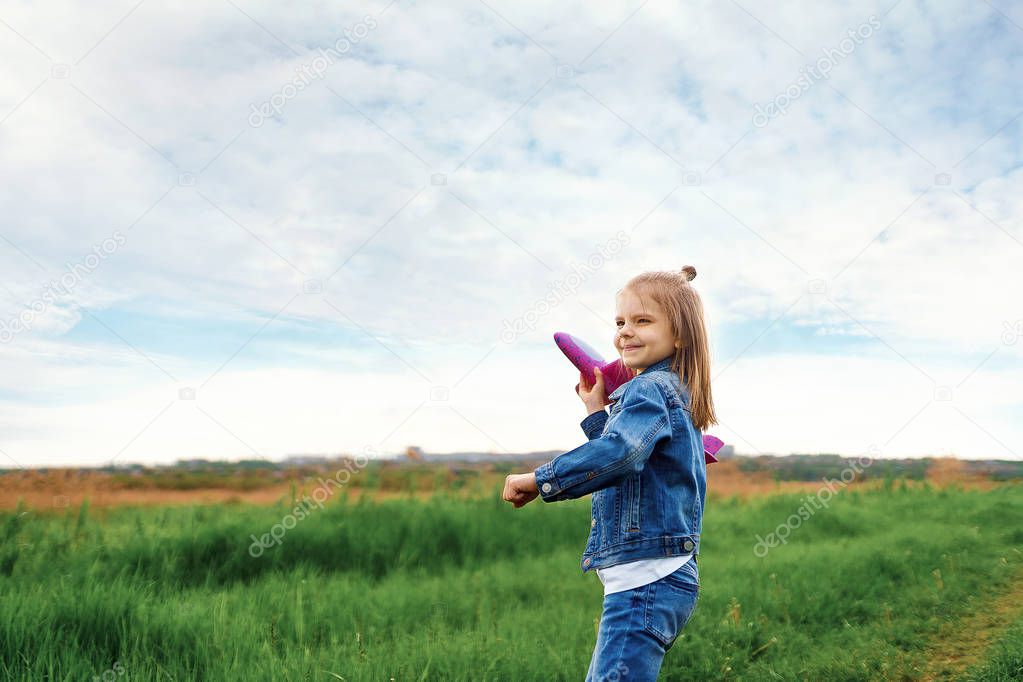 Little girl, launches a toy plane, into the air against, the background of green grass. Child launches a toy plane. Beautiful little girl, stands on the grass and launches a pink toy plane