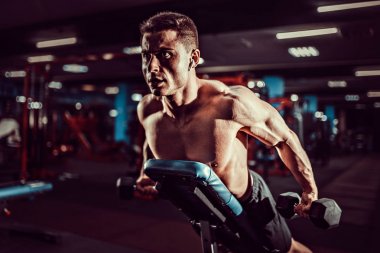 Genç kaslı adam spor salonundaki antreman bankında omuzlarının arka deltaları için dambıllarla sıkı egzersiz yapıyor.