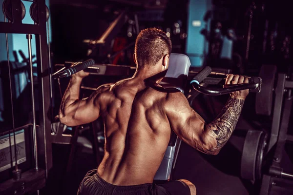 Νεαρός μυώδης άνδρας κάνει σκληρή άσκηση με αλτήρες για μέση deltas των ώμων στο μηχάνημα κατάρτισης στο γυμναστήριο — Φωτογραφία Αρχείου