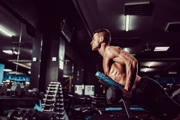Mladý svalnatý muž dělá tvrdé cvičení s činkami pro zadní delty ramen na tréninkové lavičce v tělocvičně — Stock fotografie