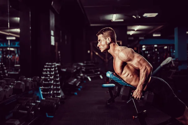 Jovem homem musculoso fazendo exercício duro com halteres para deltas traseiros de ombros no banco de treinamento no ginásio — Fotografia de Stock