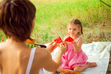 Gülümseyen iki kız çayırda açık havada karpuz dilimi yiyor. Anne ve kızı birlikte vakit geçirmek