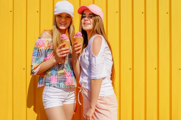 Lifestyle portret van twee mooie beste vriend hipster Lady het dragen van stijlvolle lichte outfits en het hebben van geweldige tijd. Staande in de buurt van de gele muur genieten van de dag en het eten van zoete koude ijs — Stockfoto