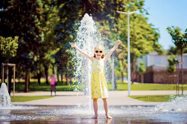 Flicka i gul klänning leker och ha kul njuter av spray av fontänen. — Stockfoto