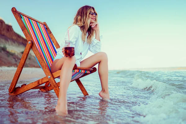 Mulher loira bonita na forma de um coração óculos de sol, camisa branca e maiô despojado relaxante em uma praia de espreguiçadeira e bebe coquetel. conceito férias de verão — Fotografia de Stock