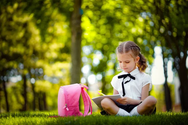 レッスンの後に草の上に座ってピンクのバックパックを持つ小さな女子高生は、本や学習のレッスンを読んで、アイデア、教育と学習の概念を考えます — ストック写真