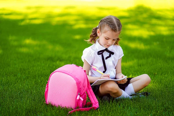 Niña de la escuela con mochila rosa sentada en la hierba después de las lecciones y leer el libro o lecciones de estudio, ideas de pensamiento, la educación y el concepto de aprendizaje —  Fotos de Stock