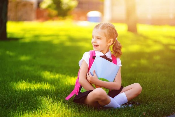 Menina da escola com mochila rosa sentado na grama depois de aulas olhando para o lado, ler livro ou estudar aulas, ideias de pensamento, educação e conceito de aprendizagem — Fotografia de Stock