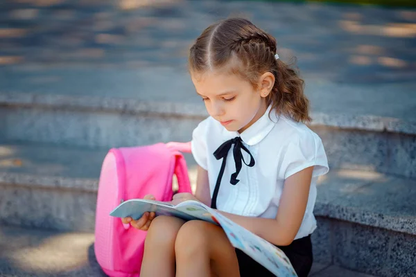 Μαθητής του δημοτικού διαβάσων βιβλίου που κάθεται στις σκάλες. Κορίτσι με σακίδιο κοντά στο κτίριο σε εξωτερικούς χώρους. Αρχή των μαθημάτων. Πρώτη μέρα του φθινοπώρου — Φωτογραφία Αρχείου