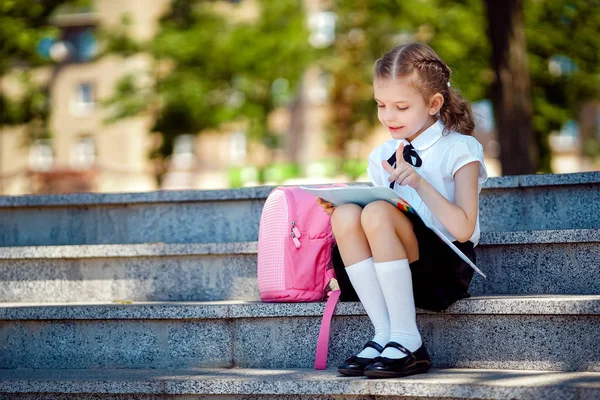 Uczeń szkoły podstawowej czytać książkę siedzi na schodach. Dziewczyna z plecakiem w pobliżu budynku na zewnątrz. Początek lekcji. Pierwszy dzień upadku — Zdjęcie stockowe