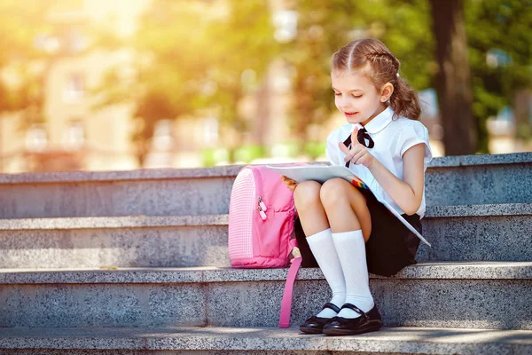 Uczeń szkoły podstawowej czytać książkę siedzi na schodach. Dziewczyna z plecakiem w pobliżu budynku na zewnątrz. Początek lekcji. Pierwszy dzień upadku — Zdjęcie stockowe