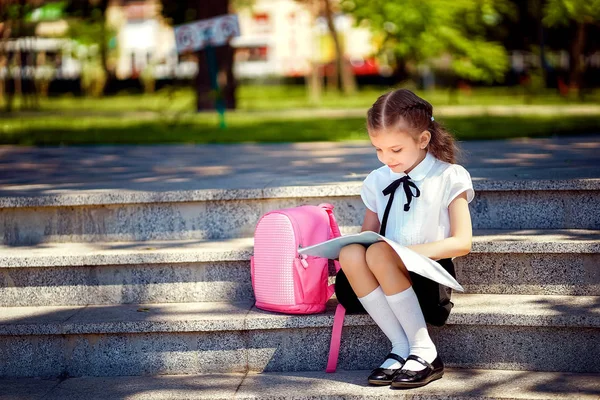 Aluno da escola primária ler livro sentado nas escadas. Menina com mochila perto de construção ao ar livre. Início das lições. Primeiro dia de queda — Fotografia de Stock