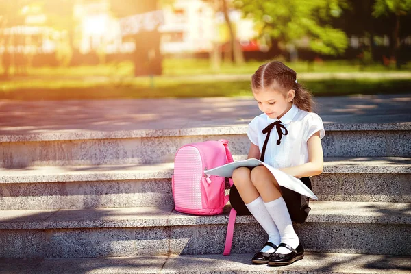 Aluno da escola primária ler livro sentado nas escadas. Menina com mochila perto de construção ao ar livre. Início das lições. Primeiro dia de queda — Fotografia de Stock